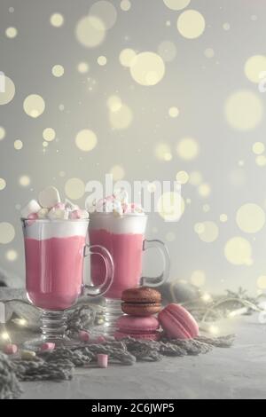 Heiße Schokolade in Rosa mit Schlagsahne und Marshmallow-Süßigkeiten in 2 Glaskrügen, französischen Makronen und weihnachtslichtern. Platz für Text kopieren. Vertikal Stockfoto