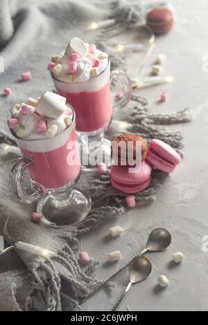 Heiße Schokolade in Rosa mit Schlagsahne und Marshmallow-Süßigkeiten in 2 Glaskrügen, süßen Pastellmakronen und Lichtern. Stockfoto