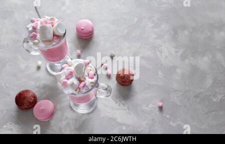 Heiße Schokolade in Rosa mit Schlagsahne und Marshmallow-Süßigkeiten in 2 Glaskrügen, süße Pastell-makronen auf grauem Hintergrund. Draufsicht Stockfoto