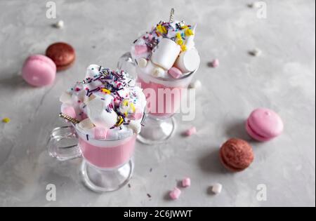 Heiße Schokolade in Rosa mit Schlagsahne und Marshmallow-Süßigkeiten in 2 Glaskrügen, süße Pastell-makronen auf grauem Hintergrund. Draufsicht Stockfoto