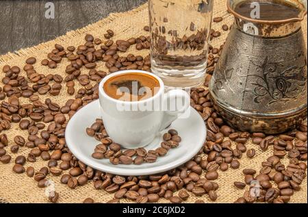 Nahaufnahme einer weißen Tasse Kaffee. Griechischer Kaffee traditionelles Konzept. Türkischer Kaffee, Bohnen, Glas Wasser und alte Kaffeekanne. Stockfoto