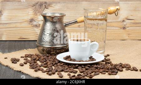 Heißer Kaffee in Türkisch Cooper und Leinentisch. Griechische Kaffeekanne, ein Glas Wasser und Kaffeebohnen fielen auf den Holztisch. Weiße kleine Kaffeetasse. Tradi Stockfoto