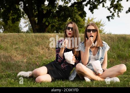 Zwei Mädchen sitzen im Park direkt auf dem Gras und essen Popcorn. Freundinnen schauen sich einen Film in einem Outdoor Kino an. Stockfoto