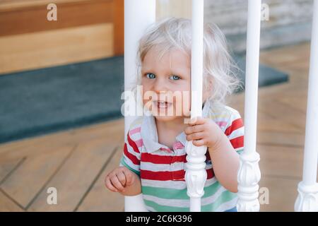 Portrait eines süßen glücklichen kleinen Jungen mit blonden Haaren und blauen Augen, hinter weißen Treppengeländern im Café im Freien stehend, wie man geht. Zurück zum normalen Konzept. Stockfoto