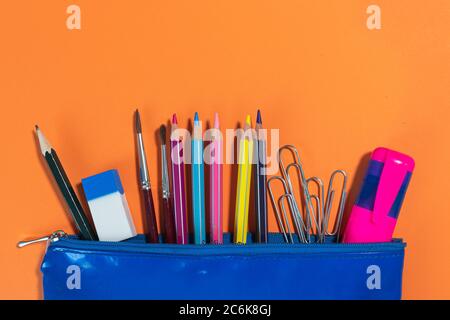 Blauer Koffer mit Schreibwaren für Schule oder kreatives Hobby auf orangefarbenem, hellem Hintergrund: Buntstifte, Pinsel, Bleistift, Clips, Textmarker Stockfoto