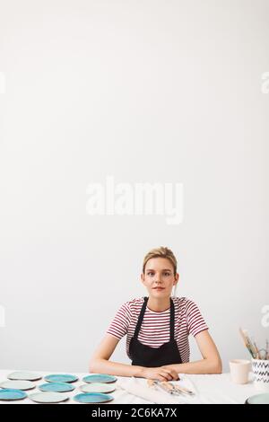 Schönes Mädchen in schwarzer Schürze und gestreiftes T-Shirt am Tisch sitzen mit handgefertigten Tellern und Tasse, traumhaft in der Kamera in Töpferei Studio Stockfoto