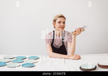 Schönes Mädchen in schwarzer Schürze und gestreiften T-Shirt sitzen am Tisch mit Tellern und hält Töpferwerkzeuge in der Hand, verträumt in der Kamera auf po Stockfoto