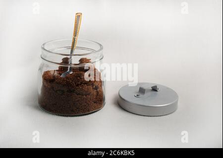 Glas mit braunem Zucker. Ein Löffel steht im Glas, der Deckel liegt neben dem Glas auf weißer Oberfläche. Stockfoto