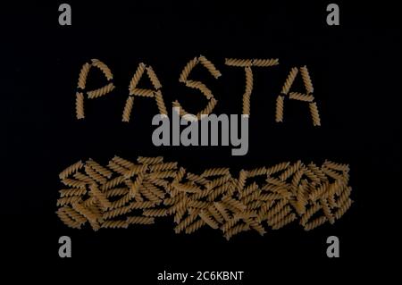 Das Wort Pasta mit Pastaschrauben geschrieben. Pasta entlang der unteren Teil des Bildes. Aufnahme mit hohem Winkel auf schwarzer Oberfläche. Stockfoto