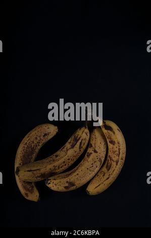 Vier überreife Bananen auf schwarzer Oberfläche. Hochwinkelaufnahme mit Kopierbereich im oberen Teil des Fotos. Stockfoto