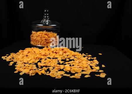 Großes Glas mit Deckel mit Maisflocken. Maisflocken verteilen sich auf schwarzem Tischtuch. Schwarzer Hintergrund mit Kopierraum im rechten Teil des Horizontes Stockfoto