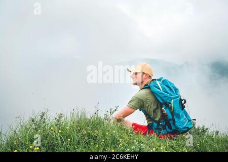 Müde Wanderer mit Rucksack Mann ruhen und genießen bewölkt Talboden Wandern durch die nebligen bewölkten Wetter Bergkette. Aktive Sportbackpacking Stockfoto