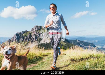 Fröhlich lächelnde Hündin beim Joggen am Wanderweg mit ihrem Beagle-Hund. Canicross Laufen gesunde Lebensweise Konzept Bild. Stockfoto