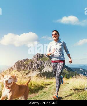 Fröhlich lächelnde Hündin beim Joggen am Wanderweg mit ihrem Beagle-Hund. Canicross Laufen gesunde Lebensweise Konzept Bild. Stockfoto