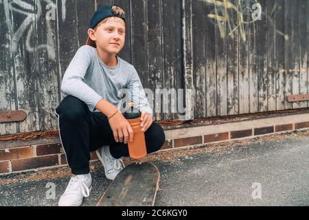 Teenager Skateboarder Junge sitzt neben einer hölzernen Grunge Graffiti Wand mit Skateboard und Wasserflasche Flasche. Jugend Generation Freetime Spending Konz Stockfoto