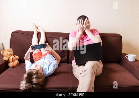 Mann, der von zu Hause aus mit einem Laptop arbeitet, mit einem kleinen Sohn, der mit dem Smartphone auf den Beinen auf der Couch liegt. Mann betonte mit Computer arbeitet zu Hause. Remote-Arbeit. Ein Stockfoto