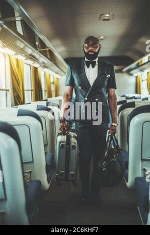 Ein stilvoller afrikanischer Unternehmer im Zug mit zwei Gepäcktaschen, die in der Mitte eines Autos stehen; schwarzer Geschäftsmann in Brille Stockfoto