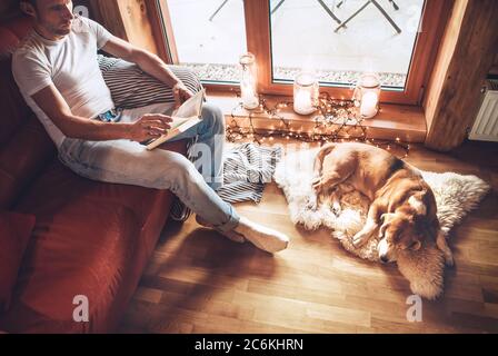 Mann liest Buch auf der gemütlichen Couch sitzen und sein Beagle Hund schläft auf Schaffell auf dem Boden. Ruhige und gemütliche Atmosphäre. Stockfoto