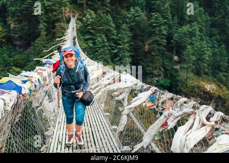 Junge lächelnde Fotografin überquert Canyon über Hängebrücke mit bunten tibetischen Gebetsfahnen geschmückt über Schlucht. Everest Stockfoto