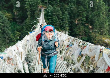 Junge lächelnde Fotografin überquert Canyon über Hängebrücke mit bunten tibetischen Gebetsfahnen geschmückt über Schlucht. Everest Stockfoto