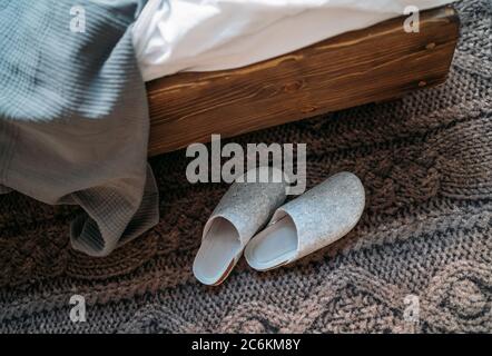 Das Paar graue Hausschuhen in der Nähe des Holzbettes auf dem "Strickboden" im gemütlichen Schlafzimmer. Home Sweet Home Konzept Bild. Stockfoto