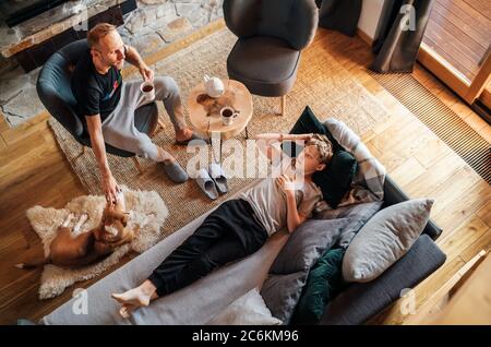 Vater und Sohn zusammen im Wohnzimmer. Junge liegt auf einem bequemen Sofa und sein Papa streichelt ihren Beagle Hund und lächelt zum Sohn. Friedliches fa Stockfoto