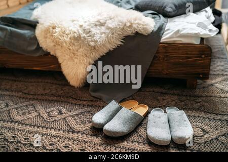 Die zwei Paar graue Hausschuhen in der Nähe des Holzbettes auf dem "gestrickten Boden" im gemütlichen Schlafzimmer. Home Sweet Home Konzept Bild. Stockfoto