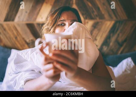 Nahaufnahme Porträt der schönen Frau sitzen auf dem weißen Leinenbett und halten die frische Kaffeetasse in den frühen Morgen. Fauler Tag frei Konzept Stockfoto