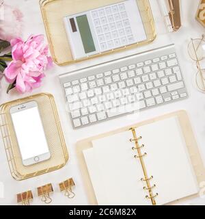 Frauentagebuch und goldene Schreibwaren. Bouquet von rosa Pfingstrosen. Brille, weiße Tastatur, Stift, Schere und Kaffee auf dem Schreibtisch. Stockfoto