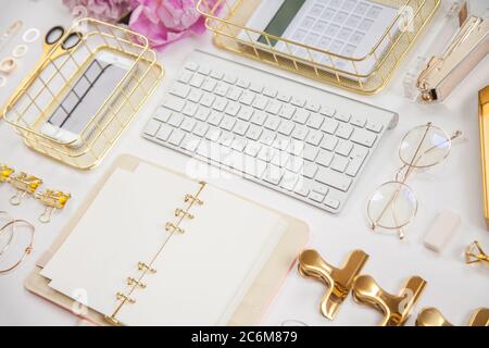 Frauentagebuch und goldene Schreibwaren. Bouquet von rosa Pfingstrosen. Brille, weiße Tastatur, Stift, Schere und Kaffee auf dem Schreibtisch. Stockfoto