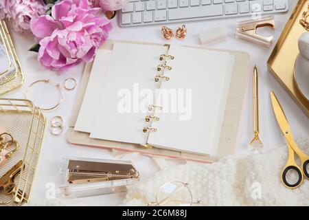 Frauentagebuch und goldene Schreibwaren. Bouquet von rosa Pfingstrosen. Brille, weiße Tastatur, Stift, Schere und Kaffee auf dem Schreibtisch. Stockfoto