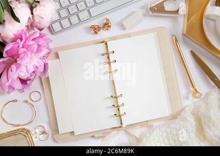 Frauentagebuch und goldene Schreibwaren. Bouquet von rosa Pfingstrosen. Brille, weiße Tastatur, Stift, Schere und Kaffee auf dem Schreibtisch. Stockfoto