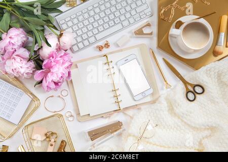 Frauentagebuch und goldene Schreibwaren. Bouquet von rosa Pfingstrosen. Brille, weiße Tastatur, Stift, Schere und Kaffee auf dem Schreibtisch. Stockfoto