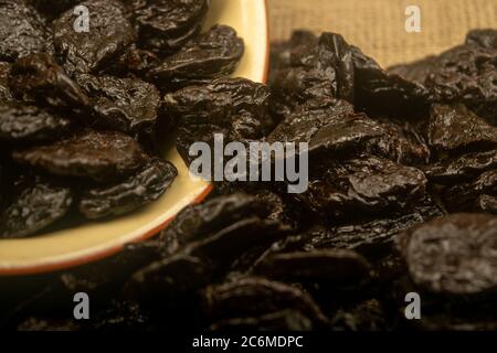 Getrocknete Pflaumen in einer Keramikschale und getrocknete Pflaumen auf einem Hintergrund von grobstrukturiertem Quappen verstreut. Nahaufnahme Stockfoto