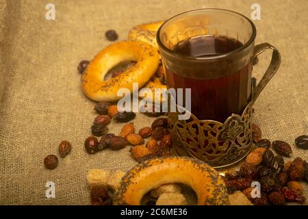 Ein geschnittenes Glas Tee in einem Vintage-Becher-Halter, Brocken von braunem Rohrzucker, kleine Bagels und getrocknete Hagebutten, die auf einem homespun Tuch mit einem Rouge verstreut sind Stockfoto