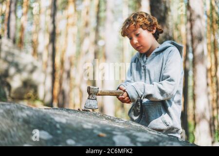 Tween boy in lässigen Outfit Schnitte Stick mit Axt auf Rock im Wald Stockfoto