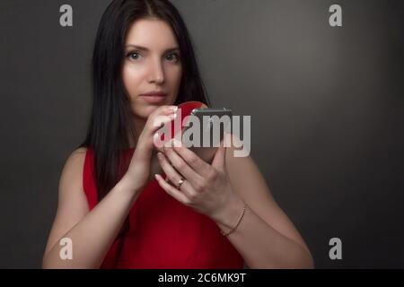 Porträt einer Brünette in einem roten Kleid. Schaut durch eine Lupe auf das Telefon Stockfoto