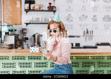 Schönes Mädchen in Sonnenbrille und Geburtstagskappe sitzt an der Theke und trinken Milchshake, während sie ihr Handy im Café. Stockfoto