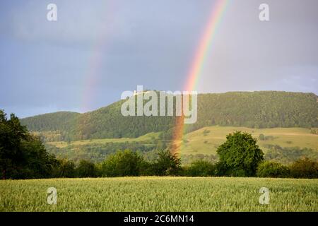 Regenbogen vor einem großen Hügel mit grünen Bäumen. Im Hintergrund sieht man das alte Schloss Teck, im Vordergrund Weizenfeld und schöne Bäume. Stockfoto