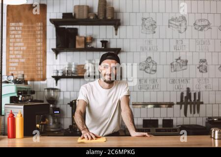 Lächelnder Barista in der Mütze stehend und wischen Bartisch, während glücklich in der Kamera im Café zu suchen. Stockfoto