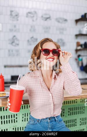 Fröhliches Mädchen in Sonnenbrille und Hemd, das an der Theke mit Sodawasser in der Hand sitzt und glücklich Zeit im Café verbringt. Stockfoto