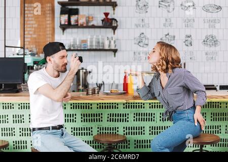 Junger Mann in schwarzer Mütze sitzt an der Theke und Fotos von schönen Dame, die ihm Luft Kuss schicken, während er Zeit im Café zusammen verbringen. Stockfoto