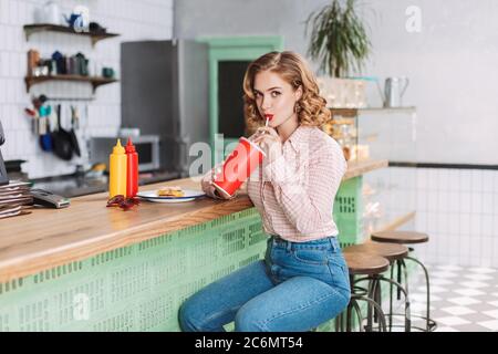 Hübsches Mädchen in Hemd und Jeans, das an der Theke im Café sitzt und Sodawasser trinkt, während es schauend in die Kamera schauend. Stockfoto