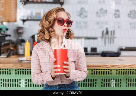 Wunderschöne Dame in Sonnenbrille und Hemd, die an der Theke sitzt und Sodawasser trinkt, während sie im Café zur Seite schaut. Stockfoto