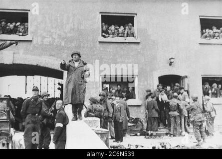 Die Übergabe des Konzentrationslagers Dachau an amerikanische Streitkräfte der 42. Infanterie-Division der Armee, 29. April 1945 in Dachau, Deutschland. US-Armee Brig. General Henning Linden, stellvertretender Generalkommandant, 42. Regenbogen-Infanterie-Division, gibt seinen Truppen Anweisungen von der Brücke zum Eingang des Jourhaus-Tores zum KZ Dachau. Der Zivilist mit dem weißen Armband ist Dr. Victor Maurer vom IKRK (Internationales Rotes Kreuz). Der deutsche Offizier links ist SS LT. Heinrich Wickert (Wicker), der deutsche Offizier, der das Lager aufgegeben hat. Amerikanische Soldaten der 7. Armee der USA, in Stockfoto