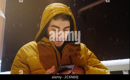 Der junge Mann im Hoodie spricht auf der Straße per Telefon, während es im Winter schneit Stockfoto