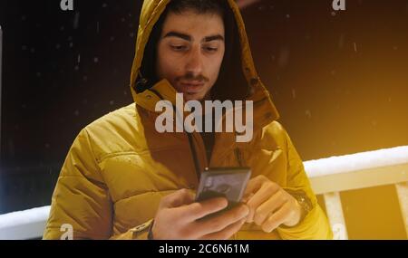 Der junge Mann im Hoodie spricht auf der Straße per Telefon, während es im Winter schneit Stockfoto