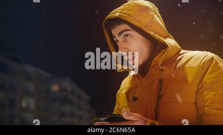 Der junge Mann im Hoodie spricht auf der Straße per Telefon, während es im Winter schneit Stockfoto