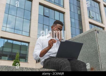 Fühlen Sie sich erschöpft. Frustriert junge schwarze afrikanische Mann die Augen geschlossen und sieht müde von zusätzlichen Arbeiten auf Laptop