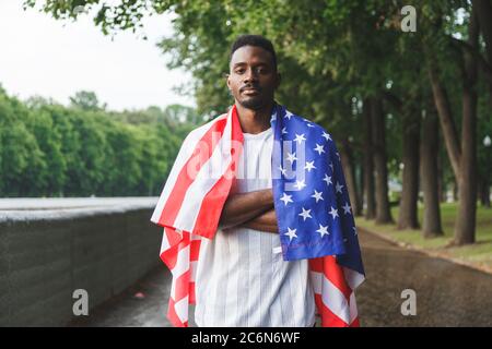 Afroamerikanischer Mann mit USA-Flagge auf den Schultern und Blick auf die Kamera, draußen stehend. Tag Sommer Stockfoto